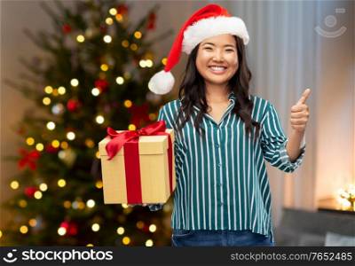 winter holidays and people concept - asian woman in santa helper&rsquo;s hat with gift box showing thumbs up over christmas tree at home on background. asian woman with christmas gift showing thumbs up