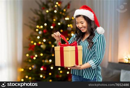 winter holidays and people concept - asian woman in santa helper&rsquo;s hat opening gift box with red bow over christmas tree at home on background. happy asian woman opening christmas gift at home