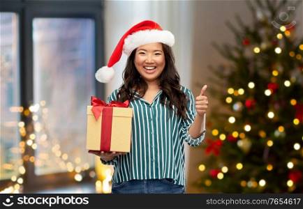 winter holidays and people concept - happy asian young woman in santa helper&rsquo;s hat with gift box showing thumbs up over christmas tree on background. asian woman with christmas gift showing thumbs up