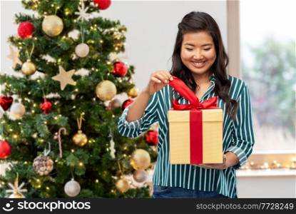 winter holidays and people concept - happy asian young woman opening gift box with red bow over christmas tree on background. happy asian woman opening gift box
