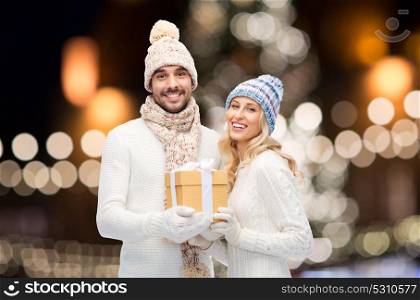 winter, holidays and people concept - happy couple with gift box over christmas tree lights background. happy couple with christmas gift over night lights