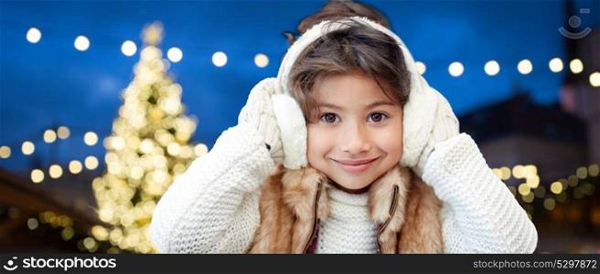 winter, holidays and people concept - happy little girl wearing earmuffs over christmas tree lights background. happy girl wearing earmuffs over christmas lights