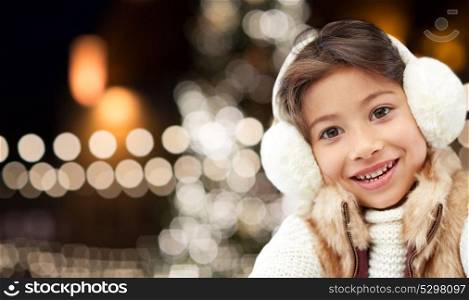 winter, holidays and people concept - happy little girl wearing earmuffs over christmas tree lights background. happy girl wearing earmuffs over christmas lights