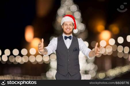 winter holidays and people concept - happy man in santa hat holding something imaginary over christmas tree lights background. happy man in santa hat holding something imaginary