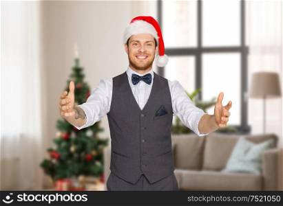 winter holidays and people concept - happy man in santa hat holding something imaginary over christmas tree on home background. happy man in santa hat holding something imaginary