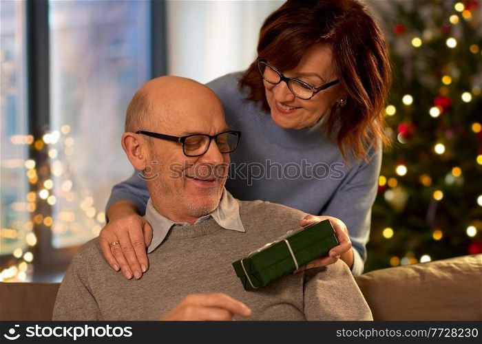 winter holidays and people concept - happy senior couple with gift box at home over christmas tree lights on background. happy senior couple with christmas gift at home