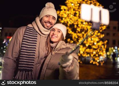 winter holidays and people concept - happy smiling couple taking picture with smartphone on selfie stick over christmas tree lights in evening city. happy couple taking selfie over christmas lights