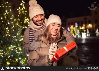 winter holidays and people concept - happy smiling couple with gift over christmas tree lights in evening city. happy couple with gift over christmas lights