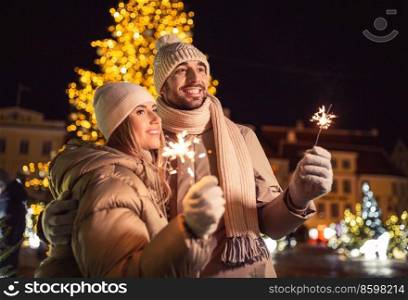 winter holidays and people concept - happy smiling couple with sparklers hugging over christmas lights in city at night. happy couple with sparklers over christmas lights