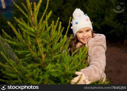 winter holidays and people concept - happy smiling little girl choosing christmas tree at street market in evening. little girl choosing christmas tree at market