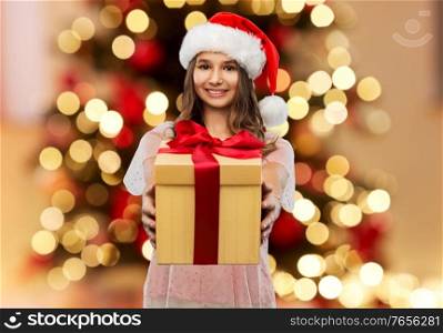 winter holidays and people concept - happy smiling teenage girl in santa helper hat holding gift box over christmas tree lights background. teenage girl in santa hat with christmas gift