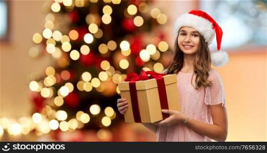 winter holidays and people concept - happy smiling teenage girl in santa helper hat holding gift box over christmas tree lights background. teenage girl in santa hat with christmas gift