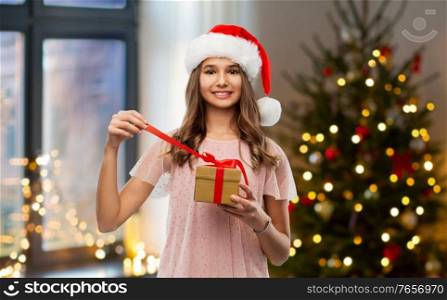 winter holidays and people concept - happy smiling teenage girl in santa helper hat opening gift box over christmas tree lights at home on background. teenage girl in santa hat opening christmas gift