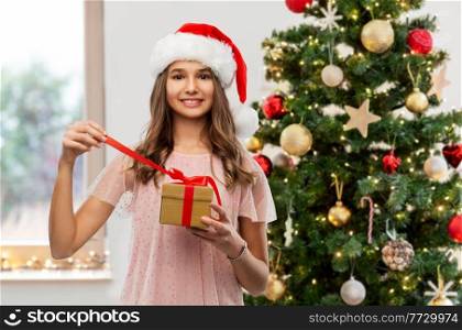winter holidays and people concept - happy smiling teenage girl in santa helper hat opening gift box over christmas tree on background. teenage girl in santa hat opening christmas gift