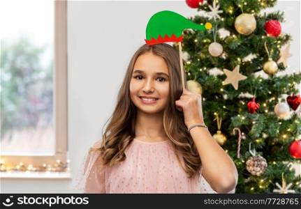winter holidays and people concept - happy smiling teenage girl with santa helper hat party accessory over christmas tree on background. happy teenage girl with santa helper hat accessory