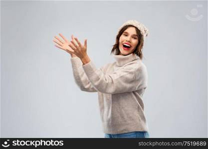 winter holidays and people concept - happy smiling young woman in hat and sweater with raised hands holding something imaginary over grey background. woman in hat and sweater holding something