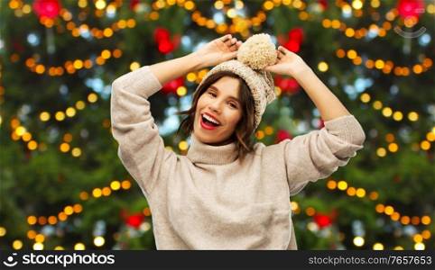 winter holidays and people concept - happy smiling young woman in knitted hat and sweater over christmas tree lights on background. happy woman in hat and sweater on christmas
