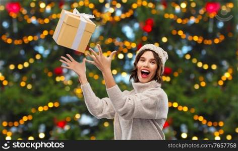 winter holidays and people concept - happy smiling young woman in knitted hat and sweater catching gift box over christmas tree lights background. young woman in winter hat catching gift box