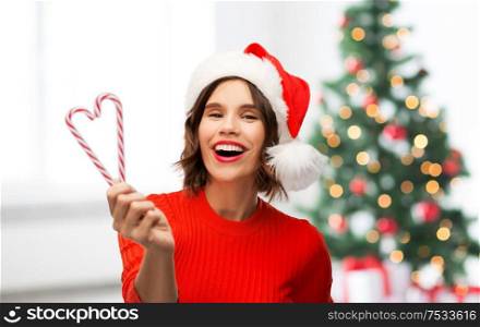 winter holidays and people concept - happy smiling young woman in santa helper hat with candy canes over christmas tree lights background. happy young woman in santa hat on christmas
