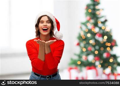 winter holidays and people concept - happy smiling young woman in santa helper hat sending air kiss over christmas tree lights background. happy young woman in santa hat sending air kiss