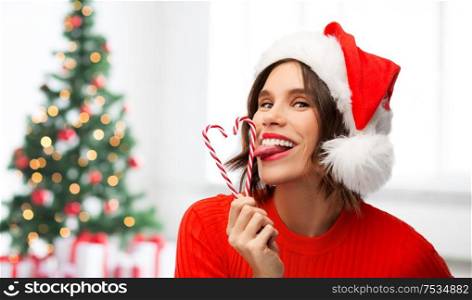 winter holidays and people concept - happy smiling young woman in santa helper hat licking candy canes over christmas tree lights background. woman in santa hat licks candy canes on christmas
