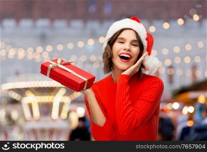 winter holidays and people concept - happy smiling young woman in santa helper hat with red gift box over christmas market lights background. happy young woman in santa hat with red gift box