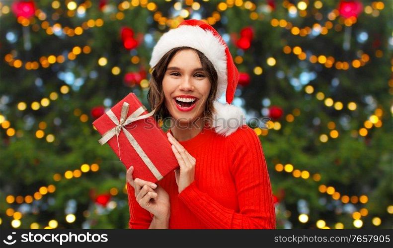 winter holidays and people concept - happy smiling young woman in santa helper hat with red gift box over christmas tree lights background. happy young woman in santa hat with red gift box