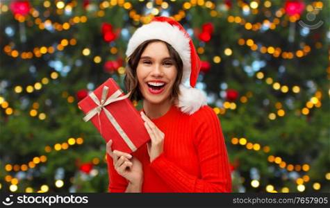 winter holidays and people concept - happy smiling young woman in santa helper hat with red gift box over christmas tree lights background. happy young woman in santa hat with red gift box