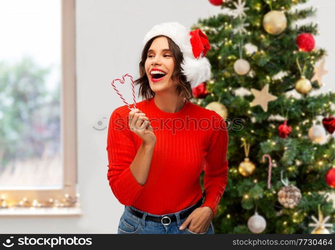 winter holidays and people concept - happy smiling young woman in santa helper hat with candy canes over christmas tree on background. happy young woman in santa hat on christmas