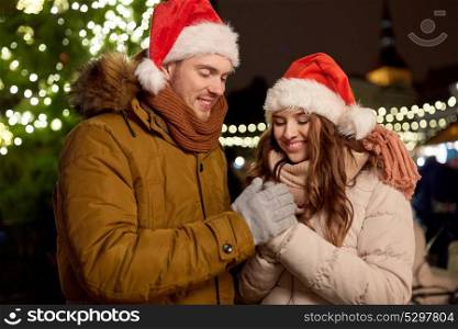 winter holidays and people concept - happy young couple in santa hats at christmas tree in evening. happy couple in santa hats at christmas tree