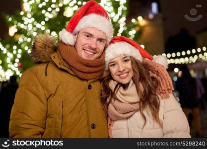 winter holidays and people concept - happy young couple in santa hats at christmas tree in evening. happy couple in santa hats at christmas tree