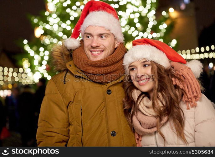 winter holidays and people concept - happy young couple in santa hats at christmas tree in evening. happy couple in santa hats at christmas tree