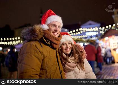 winter holidays and people concept - happy young couple in santa hats at christmas market in evening. happy couple in santa hats at christmas market