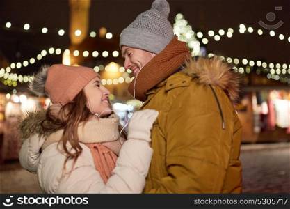 winter holidays and people concept - happy young couple with earphones dating at christmas square in evening. happy couple holding hands at christmas tree