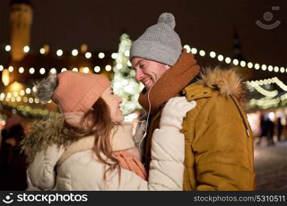 winter holidays and people concept - happy young couple with earphones dating at christmas square in evening. happy couple holding hands at christmas tree