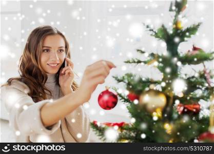 winter holidays and people concept - happy young woman decorating christmas tree and calling on smartphone at home over snow. woman with smartphone decorating christmas tree