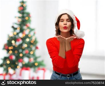winter holidays and people concept - happy young woman in santa helper hat sending air kiss over christmas tree lights background. happy young woman in santa hat sending air kiss