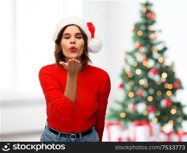 winter holidays and people concept - happy young woman in santa helper hat sending air kiss over christmas tree lights background. happy young woman in santa hat sending air kiss