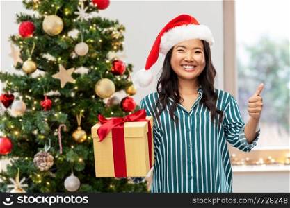 winter holidays and people concept - happy young woman with gift box showing thumbs up over christmas tree on background. happy woman with christmas gift showing thumbs up