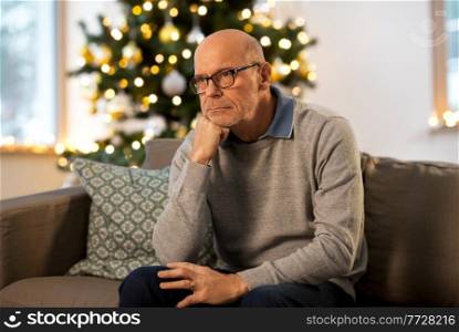 winter holidays and people concept - sad senior man in glasses thinking at home in evening over christmas tree lights on background. sad senior man thinking at home on christmas