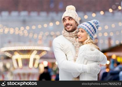 winter holidays and people concept - smiling man and woman in hats and scarf hugging over christmas market background. couple in winter hugging over christmas market