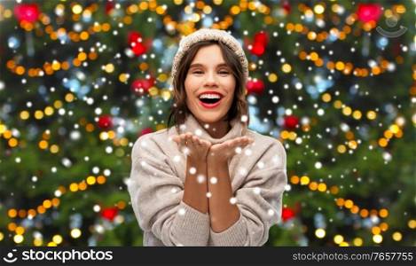 winter holidays and people concept - young woman in knitted hat and sweater holding something on palms over christmas tree lights and snow on background. happy woman over christmas tree lights and snow