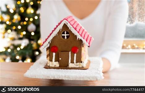 winter holidays, baking and food cooking concept - close up of woman holding gingerbread house at home over christmas tree lights background. close up of woman holding gingerbread house