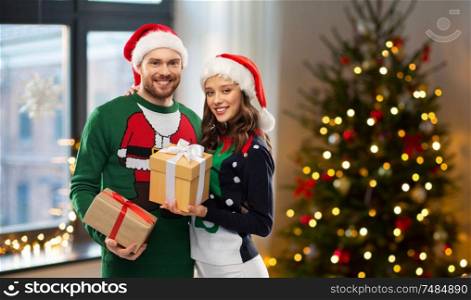 winter holidays, celebration and people concept - happy couple in santa hats and ugly sweaters with gifts at home over christmas tree lights background. happy couple in sweaters with christmas gifts