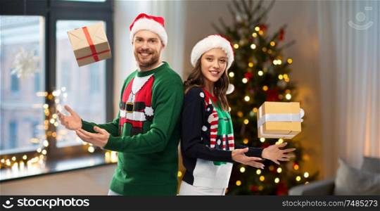 winter holidays, celebration and people concept - happy couple in santa hats and ugly sweaters with gifts at home over christmas tree lights background. happy couple in sweaters with christmas gifts