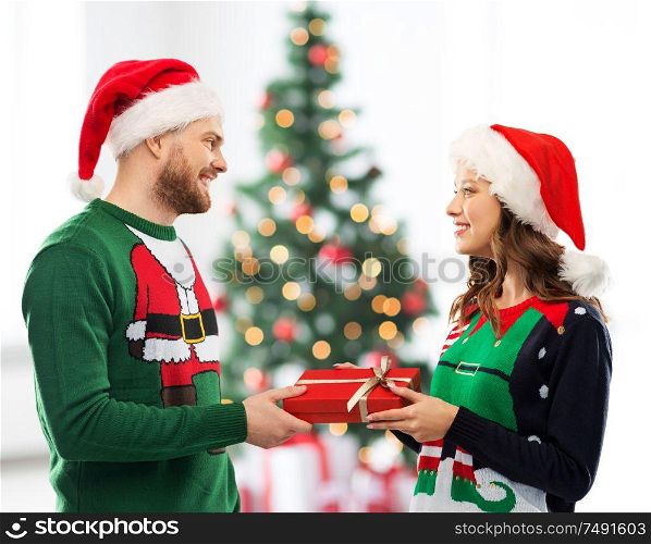 winter holidays, celebration and people concept - happy couple in santa hats and ugly sweaters with gift box at home over christmas tree lights background. happy couple in sweaters with christmas gift