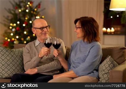 winter holidays, celebration and people concept - happy smiling senior couple toasting glasses of red wine at home in evening over christmas tree lights on background. happy senior couple drinking red wine on christmas