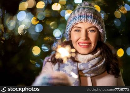 winter holidays, celebration and people concept - happy smiling woman with sparkler over christmas lights. happy woman with sparkler over christmas lights