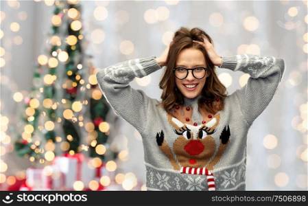 winter holidays, celebration and people concept - happy young woman wearing ugly sweater with reindeer pattern over festive christmas tree lights background. woman in christmas sweater with reindeer pattern