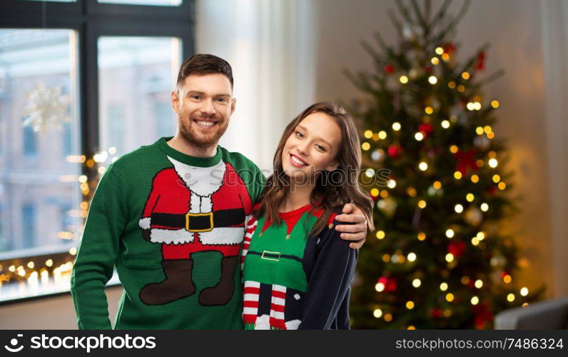 winter holidays, celebration and people concept - portrait of happy couple in ugly sweaters at home over christmas tree lights background. happy couple in ugly sweaters over christmas tree